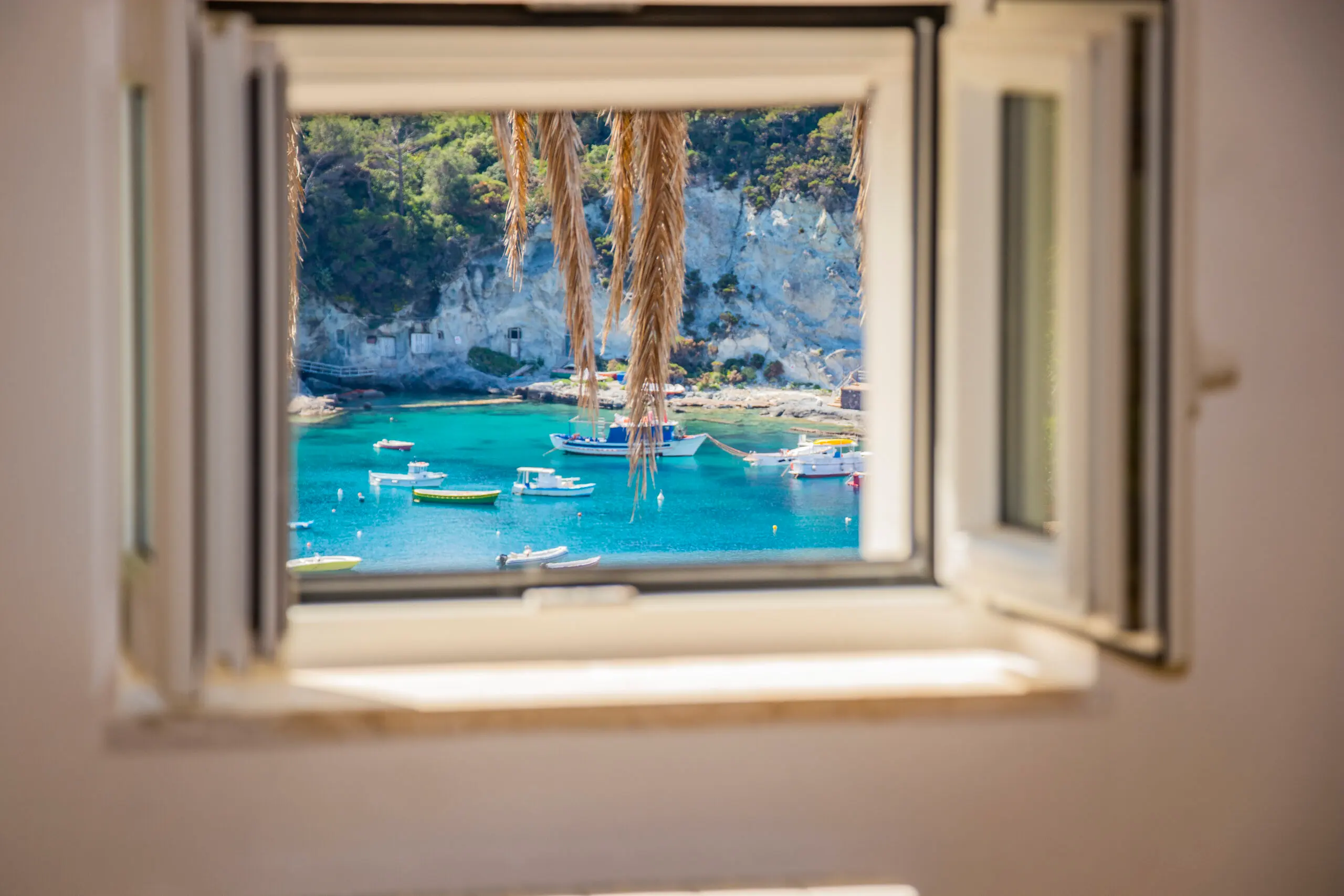Vista mozzafiato su Cala Feola a Ponza, incorniciata da una finestra aperta. Il mare cristallino e le barche ormeggiate creano un’atmosfera di relax e autenticità mediterranea.