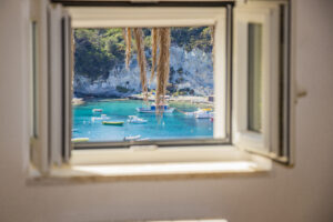 Vista mozzafiato su Cala Feola a Ponza, incorniciata da una finestra aperta. Il mare cristallino e le barche ormeggiate creano un’atmosfera di relax e autenticità mediterranea.